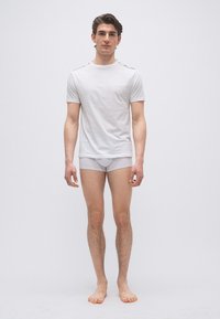 Νεαρός άνδρας, ξυπόλητος, φορώντας λευκό κοντομάνικο πουκάμισο και λευκά μποξέρ, μπροστά σε λευκό απλό φόντο.