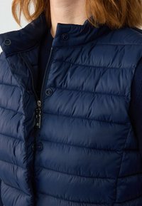 Dunkelblauer Puffer-Weste mit hohem Kragen, Reißverschluss vorne und Druckknöpfen. Verfügt über gestepptes Design, glatte Textur und metallische Reißverschlussdetails.