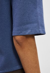 Μπλε βαμβακερό κοντομάνικο τοπ με λεία υφή και ελαφρώς φαρδιά εφαρμογή, με στρογγυλή λαιμόκοψη και τελείωμα που φτάνει λίγο πάνω από τον αγκώνα.