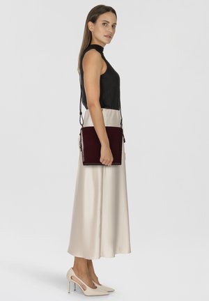 Burgundy leather crossbody bag with a sleek rectangular shape and minimal stitching. Model wears a black sleeveless top and satin cream skirt.