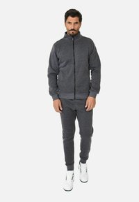 Giacca in pile grigia scura con zip e polsini a coste, abbinata a pantaloni jogger coordinati. Il modello indossa sneakers bianche. Design semplice e sportivo.