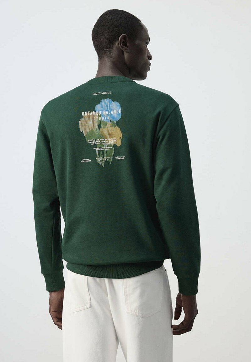 Homme portant un sweat-shirt vert foncé avec un motif abstrait coloré et du texte dans le dos, associé à un pantalon blanc, dos à un fond uni.