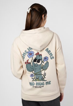 Beige hoodie featuring a cartoon cactus design with large eyes, flowers, and text "Come in my arms, no hug me" on the back. Soft fabric, relaxed fit.