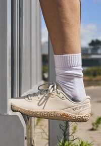 Beige Merrell shoe with a textured rubber sole, light gray laces, and a logo; worn over white crew socks on a metal railing.