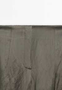 Plan rapproché du tissu de pantalon vert olive montrant la ceinture, les coutures latérales et la zone de la fermeture éclair avant.