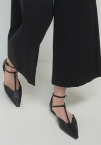 Pies que llevan planos negros de punta con tiras en T, combinados con pantalones anchos y recortados negros, sobre un suelo gris claro.