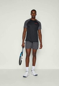 Musta urheilupaita harmailla yksityiskohdilla, lyhyet harmaat shortsit ja valkoiset urheilukengät. Pitää tennismaila kädessään. Yksinkertainen, toiminnallinen muotoilu.