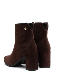 Botas de tobillo de ante marrón con punta redonda y un alto tacón cuadrado. Presentan un cierre de cremallera lateral y un pequeño acento metálico. Suela plana.