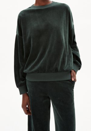 Persona vistiendo un jersey de terciopelo verde oscuro de manga larga y pantalones a juego, con una mano en el bolsillo, sobre un fondo blanco simple.