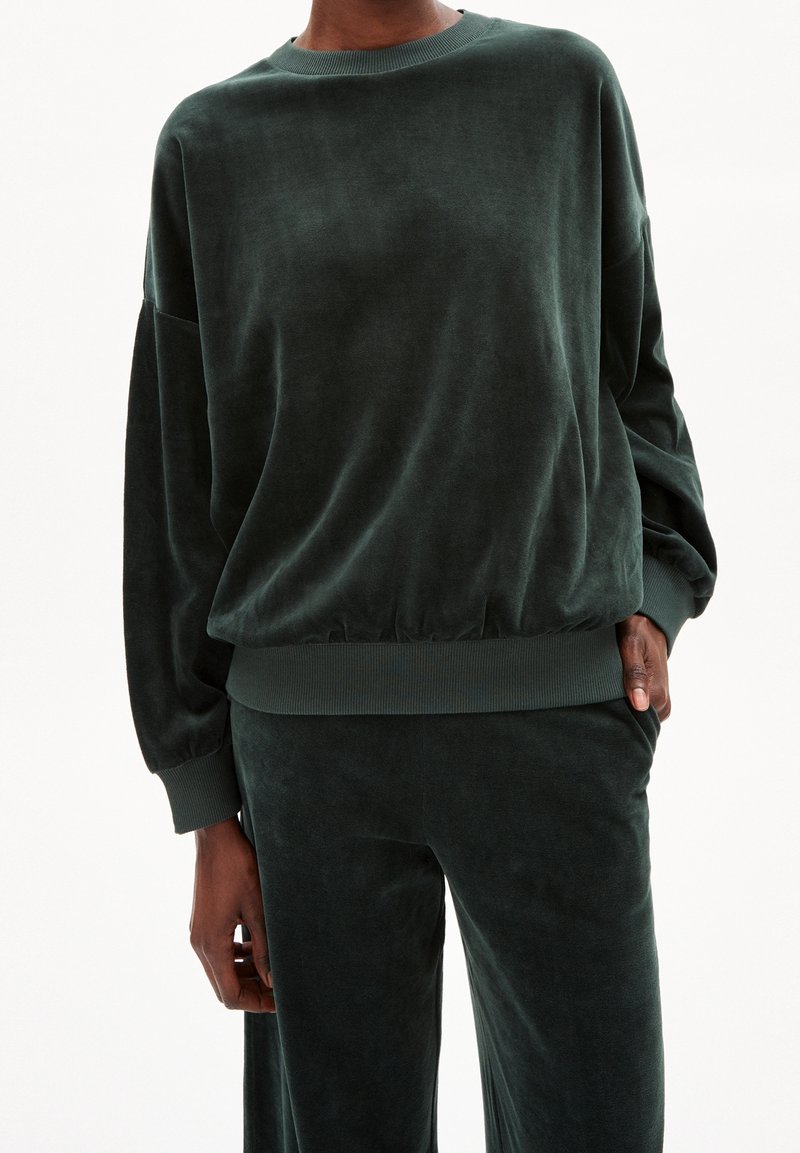 Personne portant un pull en velours vert foncé à manches longues et un pantalon assorti, une main dans la poche, sur un fond blanc uni.