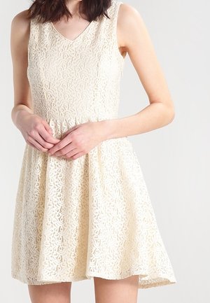 Kvinde iført ærmeløs cremefarvet blonderkjole med tætsiddende liv og udvidet nederdel, hænderne blidt foldet ved taljen.