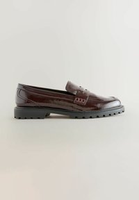 Mocassin en cuir marron foncé avec une finition brillante, une large sangle sur le cou-de-pied et une semelle en caoutchouc noir texturé. Détails subtils sur la sangle.
