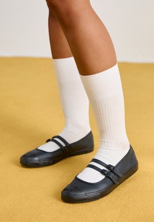 Jambes portant des chaussettes blanches côtelées hautes jusqu'aux genoux et des chaussures plates style Mary Jane noires sur un sol jaune.