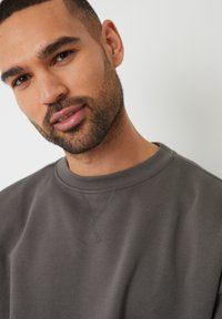 Grå sweatshirt med rund hals og subtil tekstur; har synlige syninger tæt på halsudskæringen.