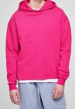 Mand iført en knaldrød hættetrøje over hvid skjorte og lyseblå jeans, stående mod en ensfarvet baggrund.
