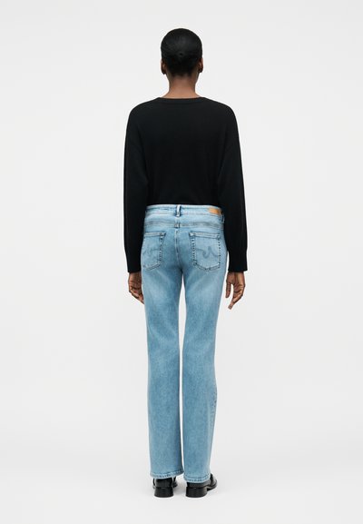 Άτομο με κοντά μαλλιά που φοράει μαύρη μπλούζα με μακριά μανίκια, ανοιχτό μπλε τζιν με ίσια εφαρμογή και μαύρα παπούτσια, όρθιο με την πλάτη γυρισμένη.