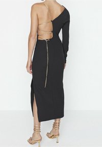 Robe noire avec une manche longue, dos ouvert orné d'accents en chaîne dorée. Présente une fente haute sur la cuisse et une silhouette ajustée.