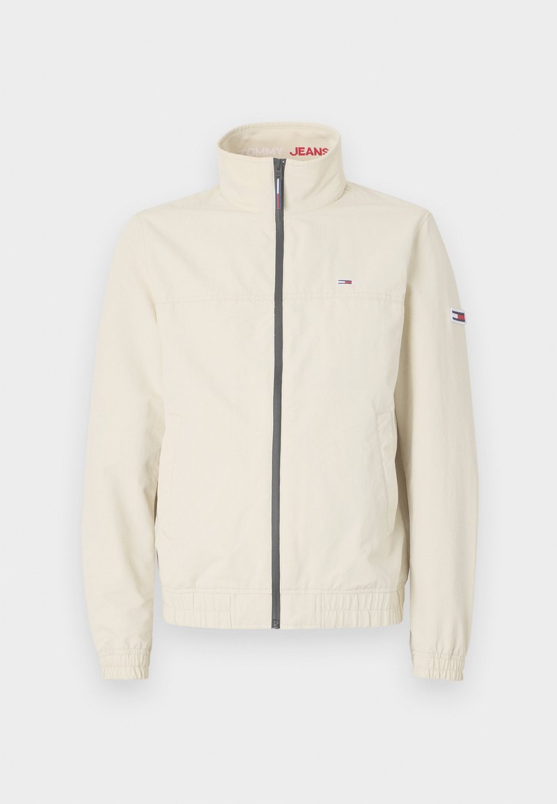 Chaqueta de color beige claro con cuello alto, cierre de cremallera, puños elásticos y bolsillos laterales. Presenta un pequeño logo en el pecho y en la manga.