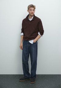 Donkerbruine trui met een hoge kraag, witte shirt eronder, losse donkerblauwe jeans en donkere schoenen. Gladde textuur en casual ontwerp.