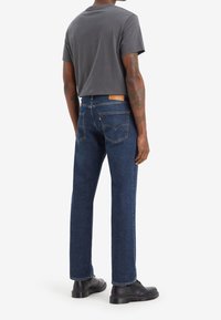 Dark blue denim jeans with a straight-leg cut, back pockets, and a brown leather patch. Paired with a gray short-sleeve T-shirt and black shoes.