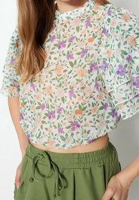 Blusa floreale con maniche corte a strati; presenta una base bianca con fiori arancioni, viola e verdi; design corto. Pantaloni corti verde oliva sotto.