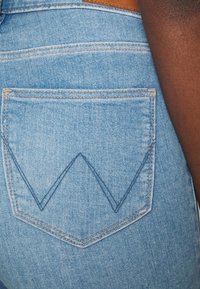 Ljust blå denimjeans med en texturerad yta, med en distinkt V-formad sömdesign på bakfickan och en brun läderetikett.