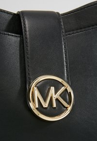 Czarna skórzana torebka z płaskim paskiem i złotym, okrągłym logo z wyraźnie widocznymi inicjałami "MK". Gładka faktura i elegancki design.