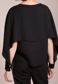 Top nero largo con un design a strati, maniche larghe e texture liscia, indossato sopra pantaloni neri aderenti con dettagli laterali.