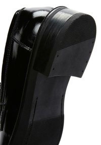 Schwarzer Lacklederschuh mit flacher, matter Sohle, die eine strukturierte Griffigkeit aufweist, sowie einem geformten Absatz, der eine glatte Oberfläche und detaillierte Nähte hervorhebt.