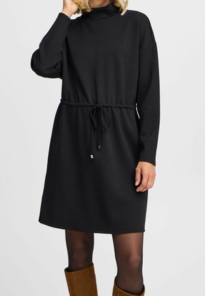 Femme portant une robe noire à manches longues avec une taille à cordon, des collants noirs transparents et des bottes en suède marron.