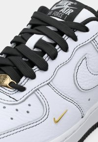 Hvit lær sneakers med svarte snørebånd, med et lite gullakksent og synlige sting. Designet inkluderer en signatur swoosh-logo.