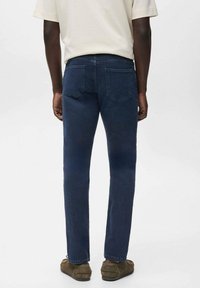 Persona vestida con pantalones vaqueros oscuros, camisa de manga corta beige y mocasines marrones, de pie con la espalda hacia una pared blanca lisa.