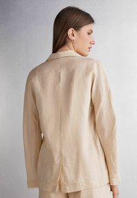 Béžový lněný blejzr s klasickým límcem, dlouhými rukávy a větracím otvorem vzadu. Textura vypadá mírně pomačkaně, což zdůrazňuje přírodní tkaninu.