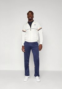 Casaco branco com fecho de correr, com riscas azul marinho e laranja, combinado com calças azul marinho e sapatilhas brancas. Apresenta um logo no peito e nos punhos.