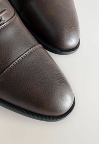Chaussures en cuir brun avec une finition texturée, bout arrondi, détails de couture subtils et semelle en caoutchouc noir. Pas d'accents matériels visibles.