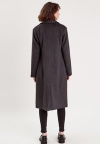 Langer, dunkelgrauer Wollmantel mit Reverskragen und minimaler Naht. Der Rücken hat eine Mittelnaht und reicht bis zur Mitte der Wade.