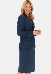Granatowy garnitur z dopasowaną marynarką z wycięciem w kształcie litery V oraz ołówkową spódnicą do kolan. Wykonany z gładkiego, teksturowanego materiału.
