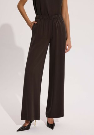 Kobieta ubrana w ciemnobrązowe spodnie o wysokim stanie i szerokich nogawkach z elastycznym pasem oraz czarne, szpiczaste wysokie obcasy, z dłońmi w kieszeniach.
