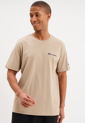 Man die lacht en een beige Champion T-shirt draagt met logo op borst en mouw, staand tegen een effen lichte achtergrond.