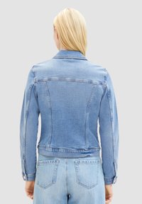 Chaqueta de mezclilla azul clara con mangas largas, cuello clásico y puños abotonados. Presenta costuras en el yugo trasero y un corte corto.