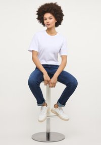 Mladá žena s afro účesem sedící na kovové stoličce, oblečená v bílém tričku, modrých zkrácených džínách a bílých teniskách.