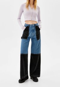 Cárdigan recortado de canalé en púrpura claro con cierre y dobladillo deshilachado, combinado con jeans de pata ancha de patchwork en denim azul y negro. Zapatillas negras de plataforma.