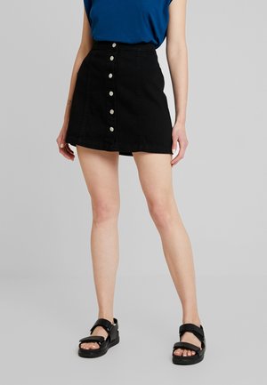 Frau trägt einen schwarzen Minirock mit Knopfleiste, ein blaues Oberteil und schwarze offene Sandalen und steht vor einem schlichten hellgrauen Hintergrund.