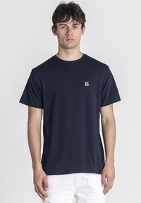 Mladý muž oblečený v jednoduchom tmavomodrom tričku s malým štvorcovým logom na hrudi a bielych nohaviciach, stojaci pred bielym pozadím.