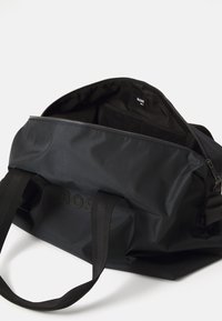 Geantă de voiaj din nylon negru, cu două mânere din material textil negre, închidere cu fermoar și un buzunar interior. Logo-ul este embosat pe partea din față.