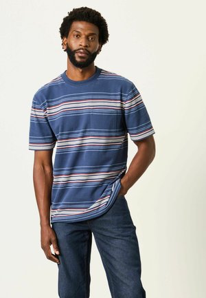 Mann mit Bart, der ein blau gestreiftes Kurzarmhemd und dunkle Jeans trägt, steht mit einer Hand in der Tasche vor einem schlichten hellen Hintergrund.
