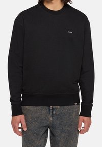 Sweat-shirt noir à manches longues avec col rond et petit logo Dickies sur la poitrine, porté avec un jean bleu délavé.