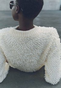 Weißer, flauschiger Pullover mit einem strukturierten, geschlungenen Design. Gerippter Ausschnitt und Ärmel. Modell von hinten gezeigt, mit dunkler Sonnenbrille.