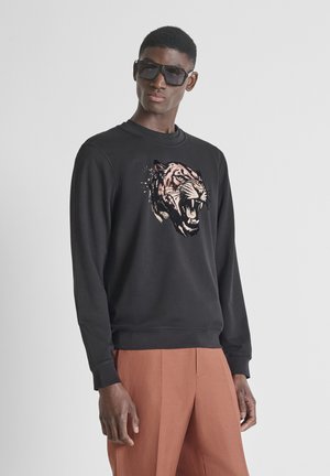 Uomo con felpa nera con grafica di tigre ruggente, pantaloni marroni e grandi occhiali da sole neri, in piedi davanti a uno sfondo chiaro e uniforme.