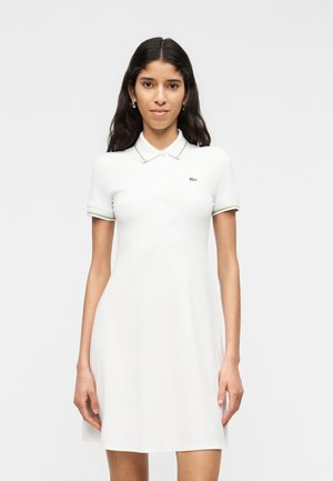 Mujer con cabello largo y oscuro, vestida con un vestido polo blanco de manga corta con cuello y bordes de las mangas en verde, de pie contra un fondo uniforme.